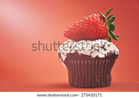 Close-up of a delicious red strawberry on a mini chocolate muffin cupcake against a red orange background, with copy space and applied retro style filters and added lens flare light beam.