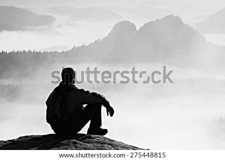 Hiker in black on the rocky peak. Wonderful daybreak in mountains, heavy orange mist in deep valley. Man sit on the rock. Black and white photo.