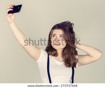 photography. Cheerful young  woman holding mobile phone and making photo of herself while standing against grey background