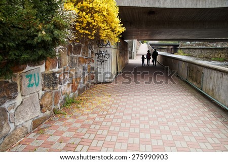 The paved walkway that runs along the waterfront along the river. Silhouettes of pedestrians walking under the bridge. Flowering shrubs over a stone wall.