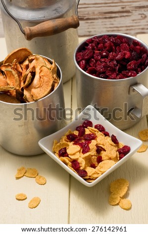 Vertical photo of white square bowl full of cornflakes with dried cranberries. Few flakes are around and two aluminum cups full of fruit with one small can in background. 