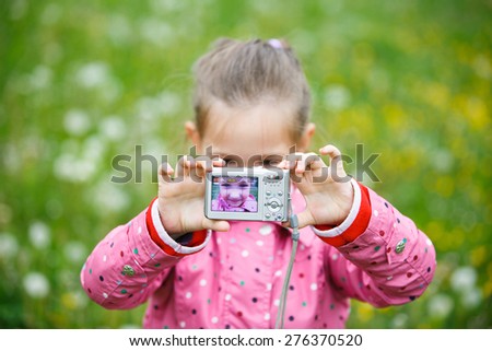 Little cheerful girl making a selfie with digital camera, enjoying her time on a dandelion meadow. Active lifestyle, curiosity, pursuing a hobby, technology and kids  concept.  