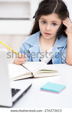 Funny picture of little dark-haired girl playing role of business woman. Girl sitting at table with laptop, writing at notebook and using phone. Office interior as a background