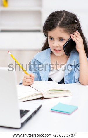 Funny picture of little dark-haired girl playing role of business woman. Girl sitting at table, writing at notebook and using phone. Office interior as a background