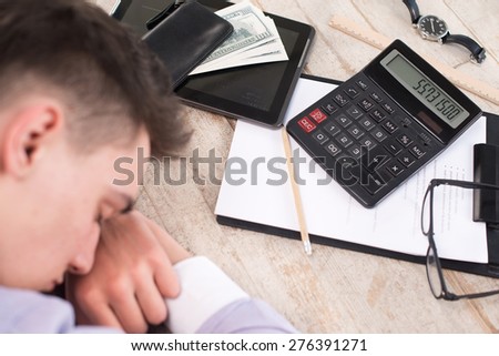 Creative top view photo of young businessman sleeping near paper, watches, purse with money, pencil, calculator, straight edge tool, glasses and tablet computer on the light-colored woodblocks