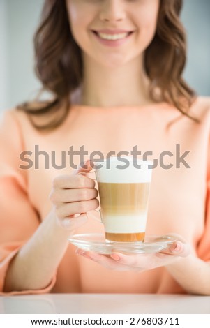 Close-up  of smiling girl in peach blouse  holding glass of coffee with cream on grey background.