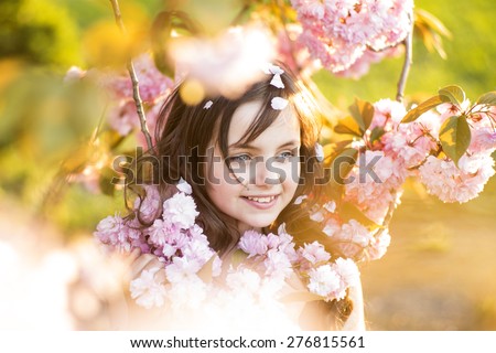 Small brunette girl looking away standing amid pink japanese cherry blooming in broad daylight in the park, horizontal picture