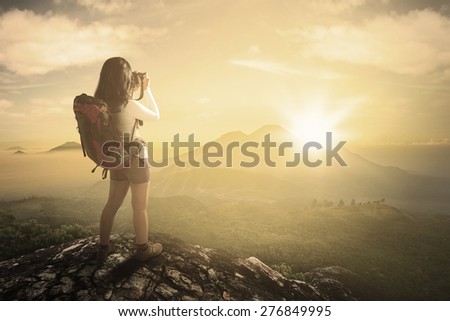 Rear view of young woman standing on the mountain while carrying backpack and taking picture by using digital camera