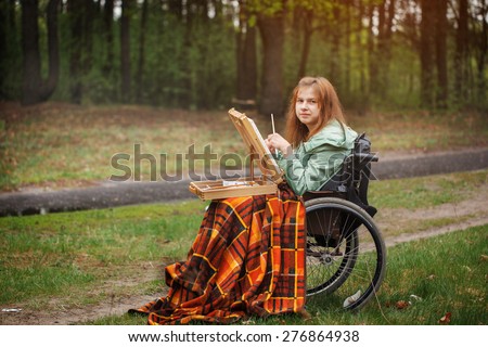 Young smiling woman in a wheelchair covered by a checkered plaid.Young girl paints   picture in spring forest.