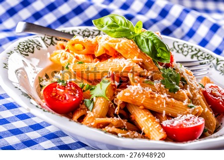 Plate with pasta pene Bolognese sauce cherry tomatoes parsley top and basil leaves on checkered blue tablecloth. 