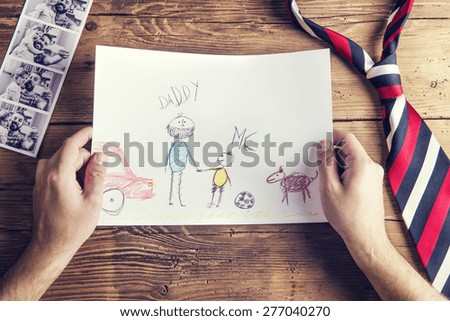Pictures of father and daughter, childs drawing and tie laid on wooden desk backround.