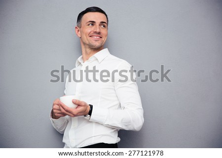 Happy young businessman holding cup of coffee and looking way at copyspace. Standing over gray background