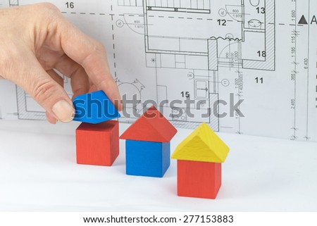 Three houses made of wooden multicolored shapes are standing in front of the blueprint of the house. Female hand is giving a blue roof on one of the homes.