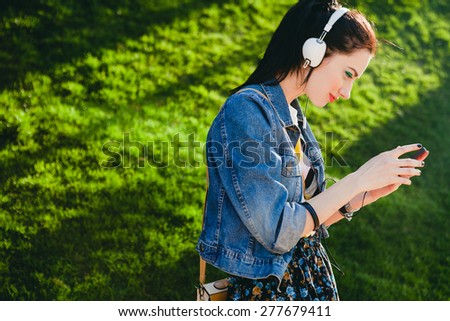 young hipster stylish beautiful girl listening to music, mobile phone, headphones, enjoying, denim outfit, smiling, happy, cool accessories, vintage style, having fun, looking, park