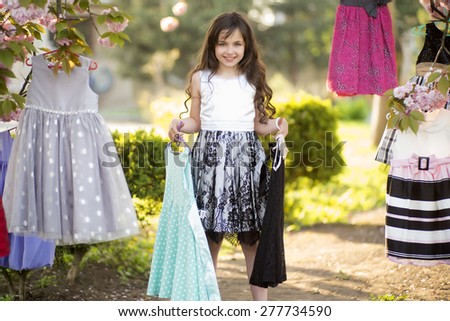 Little cute girl looking forward choosing colorful baby clothes in the cherry bloom pink flowers tree in broad daylight in the park, horizontal picture