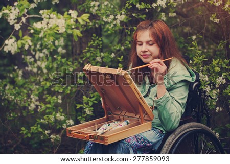 Portrait young woman sitting in a wheelchair , smiling and painting on small easel. Toned image

