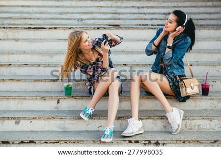 two young beautiful happy stylish hipster girls, cocktail drink, denim outfit, smiling, happy, cool accessories, vintage style, having fun, sitting, stairs, sneakers, headphones, camera, take photo