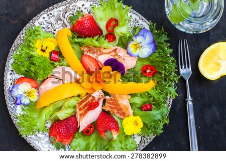 Salad with smoked salmon, strawberries and edible flower on black wooden background, top view