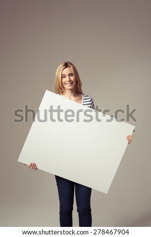 Smiling Pretty Blond Girl Holding an Empty White Sign Board, Emphasizing Copy Space, and Looking at the Camera. Captured in Studio with Brown Background.