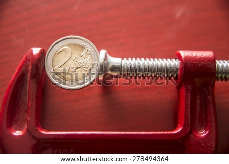 two euro in the clamp coin of 2 euro tightened up in a G-clamp, financial crisis concept, idea of  inflation on red wooden background. Royalty-Free Stock Photo #278494364