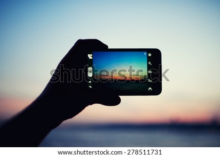 Person taking a photo of amazing sunset using smart phone camera, tourist hand holding cell phone while taking a photograph of nature landscape in travel,taking a picture of outdoors, blur background