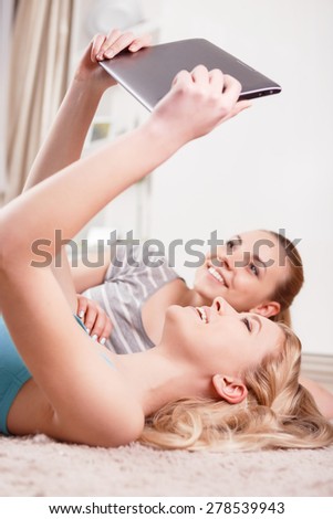 Friendship. Two young beautiful blond girls wearing pajamas lying on the floor holding a tablet above and looking at it smiling side view 