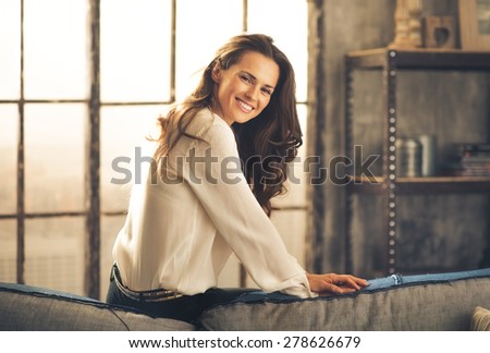 An elegant brunette sitting on the back of sofa in a loft, leaning forward and smiling is looking over her shoulder. In the background, the daylight is streaming through a window. Chic loft.