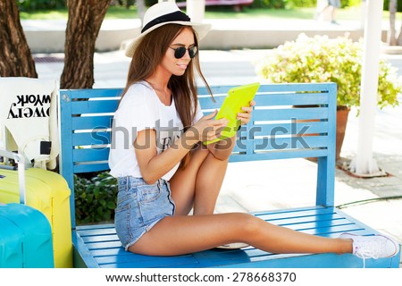 young girl works on the tablet on the internet and goes to the airport.working on tablet in  park,using interenet connection.Summer image,wear hat and summer sunglasses,bright luggage cool accessories