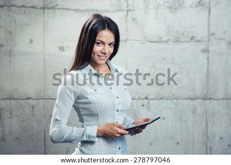 Portrait of a cheerful businesswoman with tablet computer standing over concrete wall and looking at camera