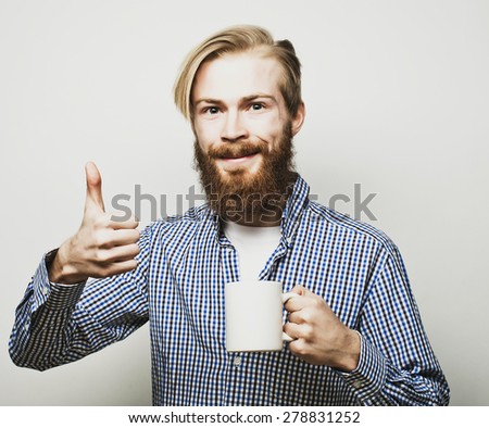 life style, happiness and people concept: young bearded man with a cup of coffee in hand and showing okey, against grey background.