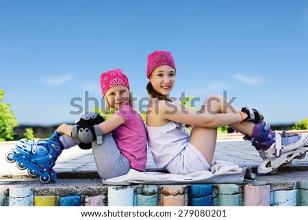 
Two girls in roller skates sitting side by side on the street.