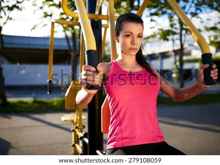 Woman exercising her upper body using weights machine outside and looking at camera.