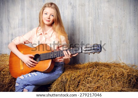 Beautiful girl teenager in shirt and torn jeans playing guitar sitting on hay. Jeans fashion. Western style.