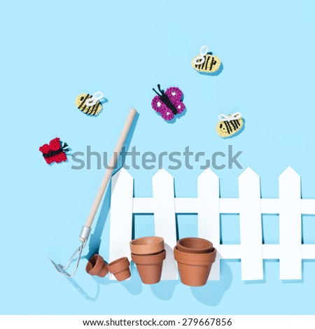 crochet butterflies and bees with a white paper fence, pots and a rake on blue background