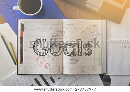 Notebook with text inside Goals on table with coffee, some diagrams on paper and laptop 