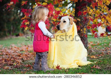 A cute little toddler girl playing with her dog, a yellow labrador in the park with autumn trees in the background