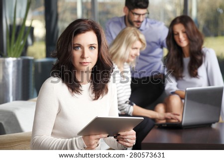 Image of mature businesswoman using her digital tablet while sitting at office and business people consulting at background with laptop. 
