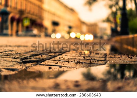 City central square paved with stone after a rain, headlights from cars in the distance. View from the pavement level next to the roadside puddle, image in the yellow toning