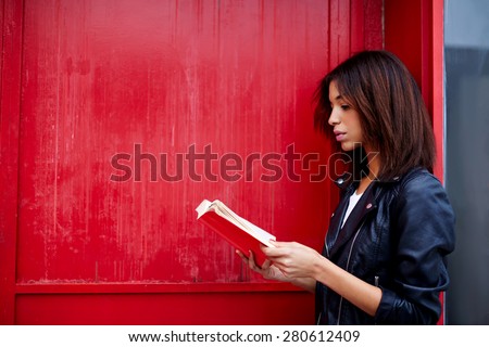 Young female student reading interesting book while standing in the city on red wall background with copy space for your text message, afro american woman read literature while standing outdoors