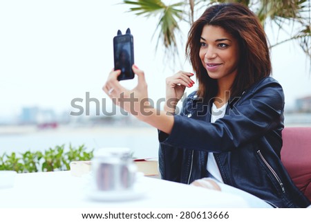 Gorgeous young woman making self portrait with a cell phone camera while enjoying a day out and sitting in coffee shop at her recreation time, female hipster taking a picture of herself on smart phone