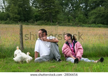 Man and young woman together, sitting in a field with baby ducklings next to the man.