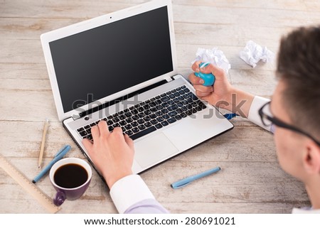 Creative top view photo of young businessman using anti-stress ball while working with laptop. Man sitting near cup of coffee, straight edge tool, crumpled paper on the light-colored woodblocks