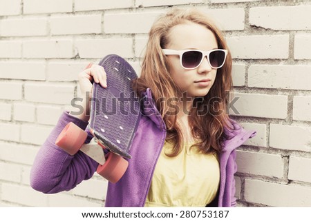 Blond teenage girl in sunglasses holds skateboard near gray urban brick wall, vintage tonal correction, old style photo filter effect