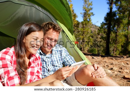 Camping couple in tent using smartphone or small tablet looking at pictures photos. Campers smiling happy outdoors in forest. Happy multiracial couple having fun outdoor. Asian woman, Caucasian man