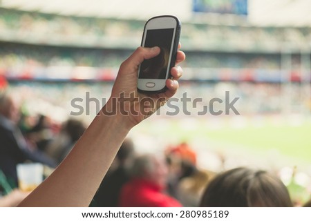 A female hand is holding a smart phone in a stadium to take pictures of a sporting event