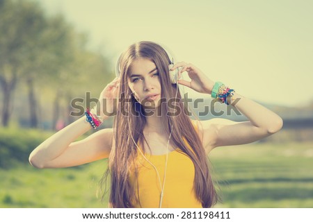 Beautiful teenage girl listening to music on headphones in park. Closeup of attractive young blonde Caucasian woman in yellow tank top with white headphones outdoors in summer. Color filter applied.