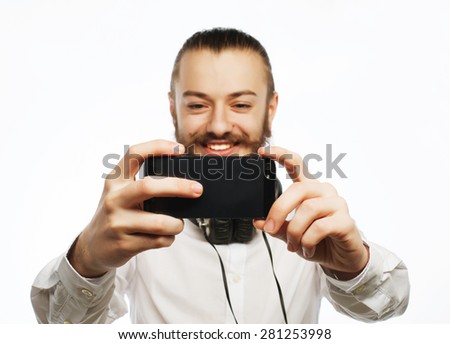 Internet, tehnology and people concept: a young man with a beard in shirt holding mobile phone and making photo of himself while standing against white  background.