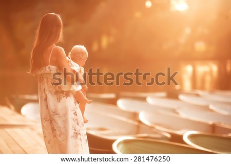 beautiful mother is standing on the pier and holding her little baby girl daughter on her hands and hugging her very gently at summer time on sunset