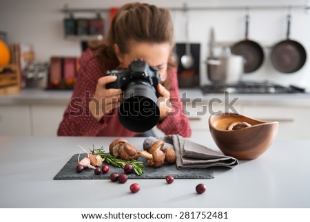 A woman food photographer in the background leans down to take a close-up, in a modern kitchen, of autumn fruits and vegetables - mushrooms, garlic, rosemary, and cranberries. Royalty-Free Stock Photo #281752481