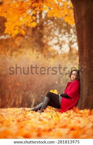 beautiful pregnant woman in red coat is sitting under the yellow tree in the autumn park